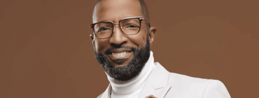 Man in glasses wearing white suit smiling.